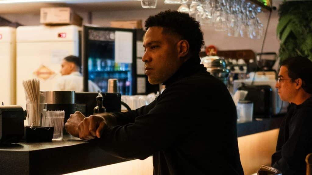A man in a dark turtleneck sits at a bar counter, looking off to the side.