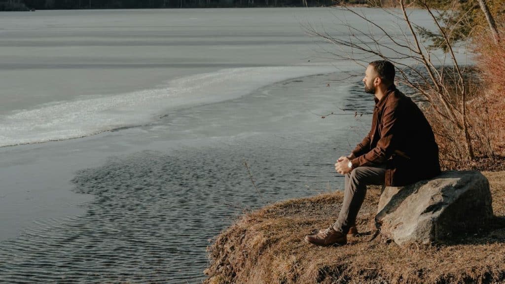 A bearded man sits on a rock by the shore of a partially frozen lake, looking out at the water.