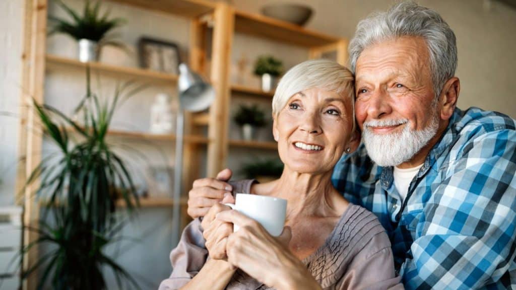 A happy elderly couple, with the man embracing the woman holding a mug, smiles in a cozy room.