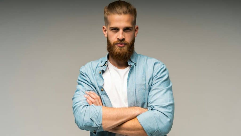 A serious bearded man in a blue shirt and white tee stands with arms crossed.
