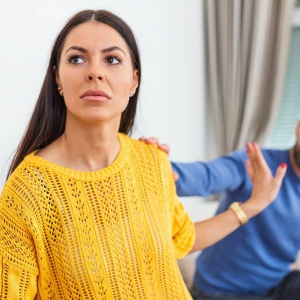 A woman in a yellow sweater looks away while a man behind her reaches out.