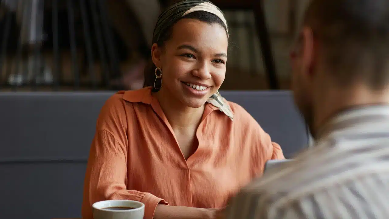 A smiling woman talking with another person over coffee in a cozy café.