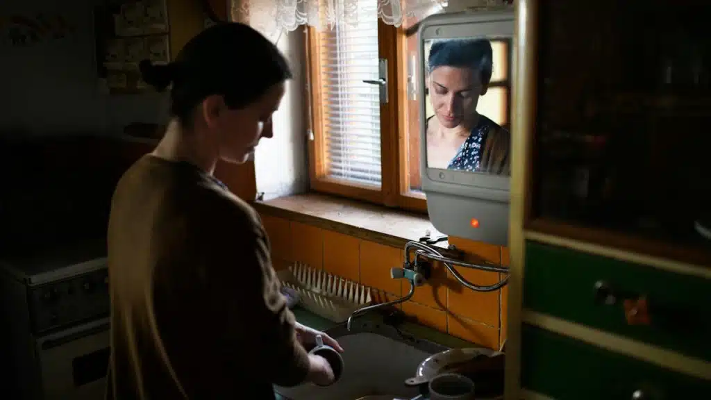A woman washes dishes in a rustic kitchen, her contemplative expression captured in a small mirror reflection above the sink.