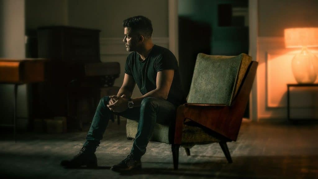 A man sits alone in an armchair in a dimly lit room, looking away thoughtfully.