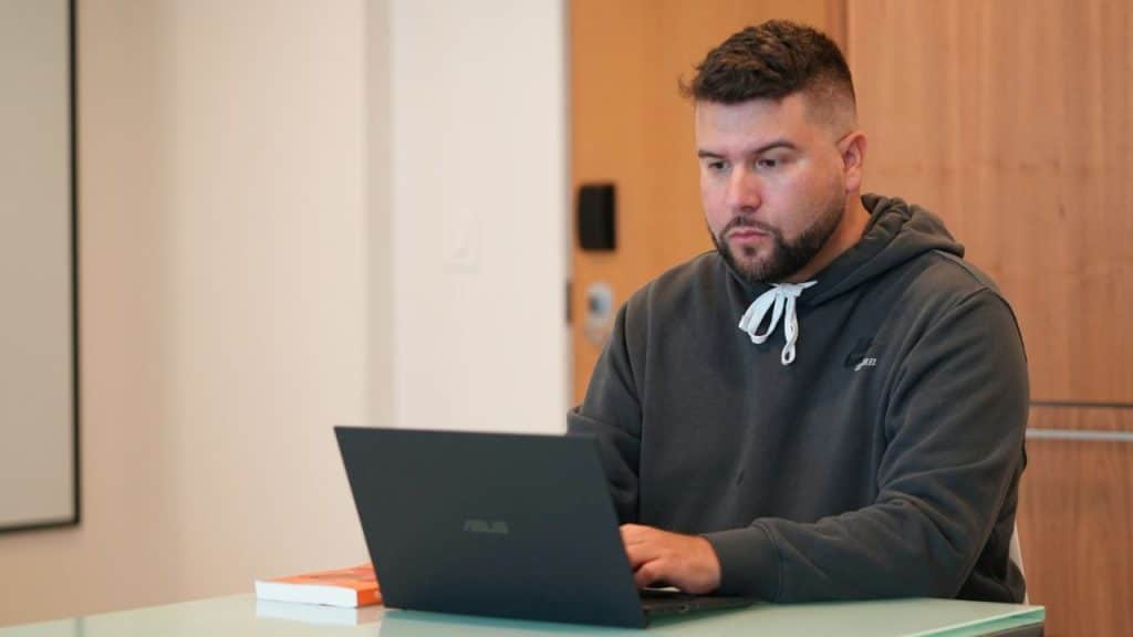 A man in a grey hoodie sitting at a table and using a laptop.