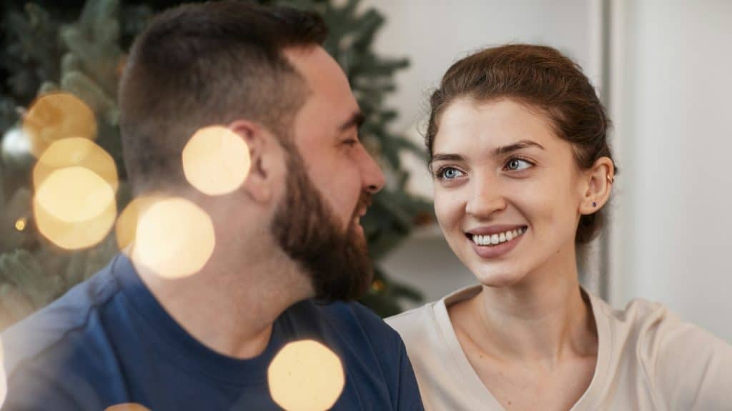A man with a beard and a woman smile while looking at each other indoors.