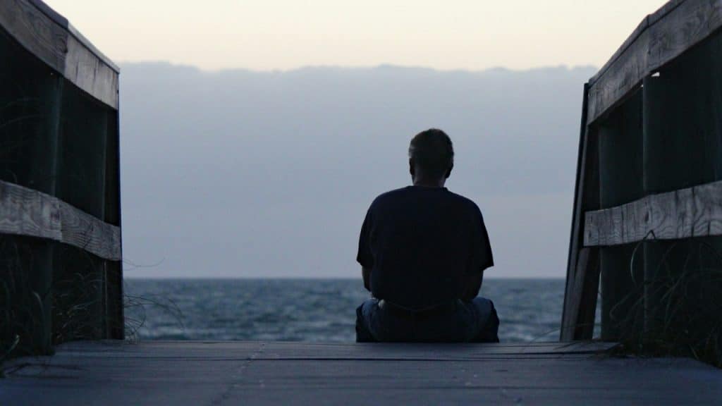 Silhouetted view of a man sitting on a wooden boardwalk facing the ocean at dusk.