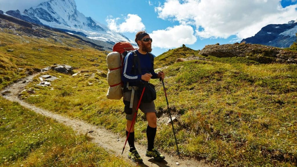 A bearded man with a large backpack and trekking poles hikes a mountain trail.