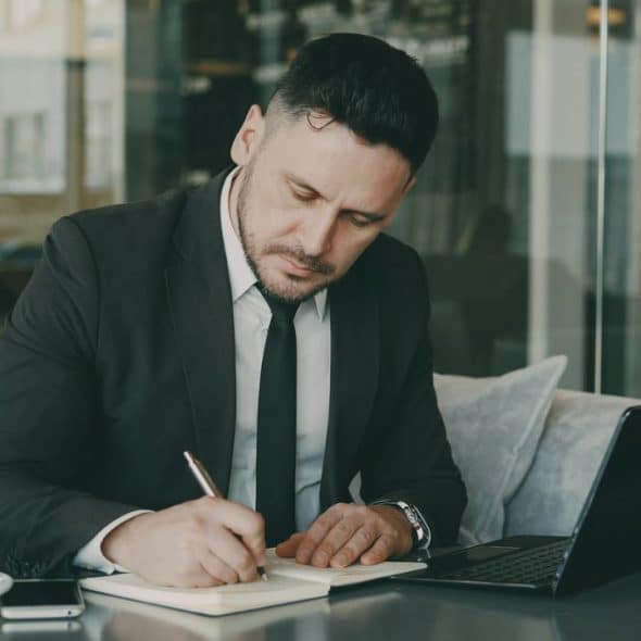 A man in a suit sits at a table writing in a notebook near a laptop.