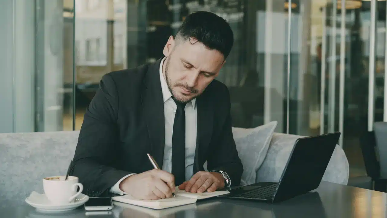 A man in a suit sits at a table writing in a notebook near a laptop.