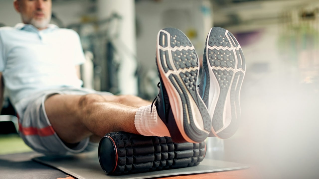 A person sits on a mat using a black foam roller under their lower legs.