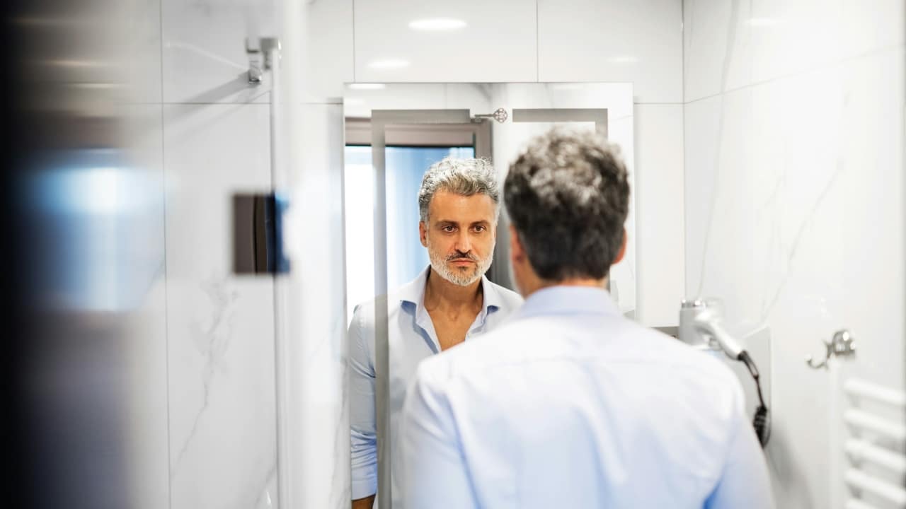 A man with gray hair looks at his own reflection in a bathroom mirror.