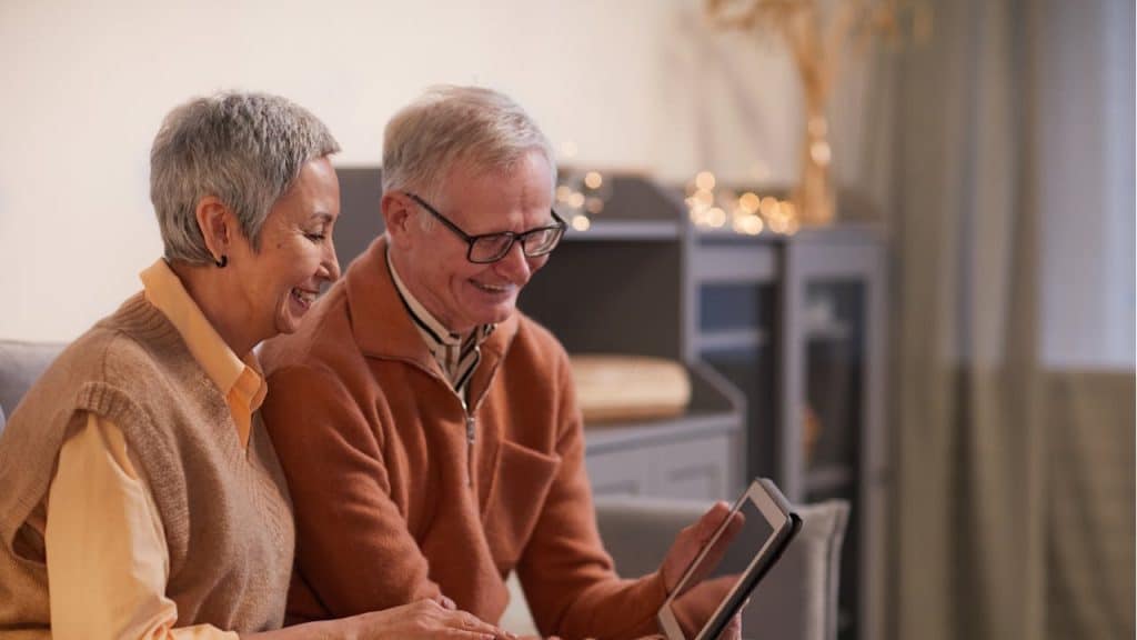 A smiling older man and woman sit together on a couch while looking at a tablet in a cozy, well-lit room.