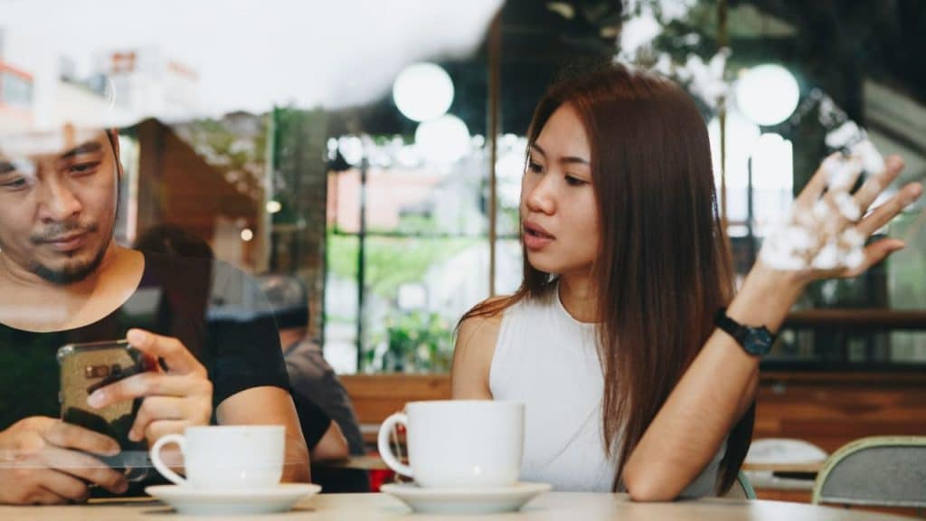A man looks at his phone in a cafe while a woman gestures and speaks.