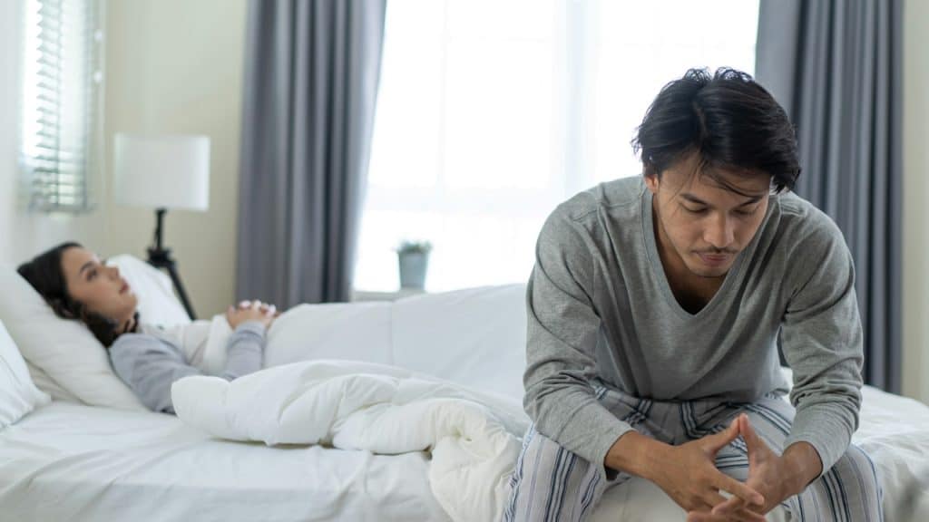 A man sitting on the bed looking anxious and clasping his hands while a woman lies in the background.