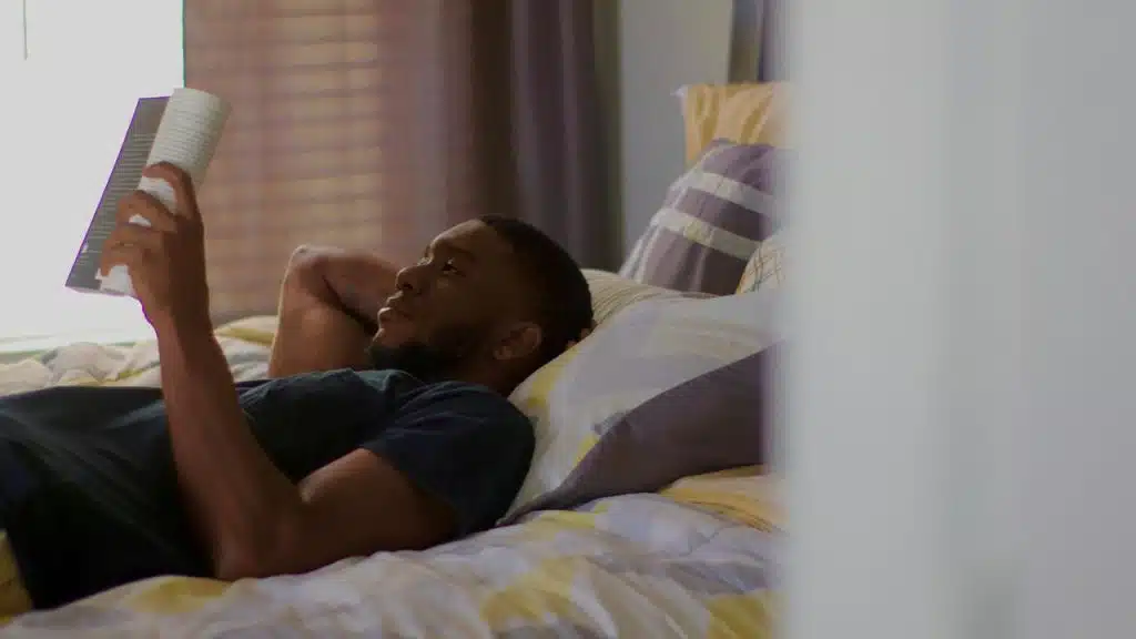 A man lying on a bed reading a book in soft light.