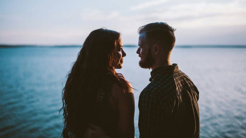 A man and woman stand in silhouette, facing each other in front of a body of water.