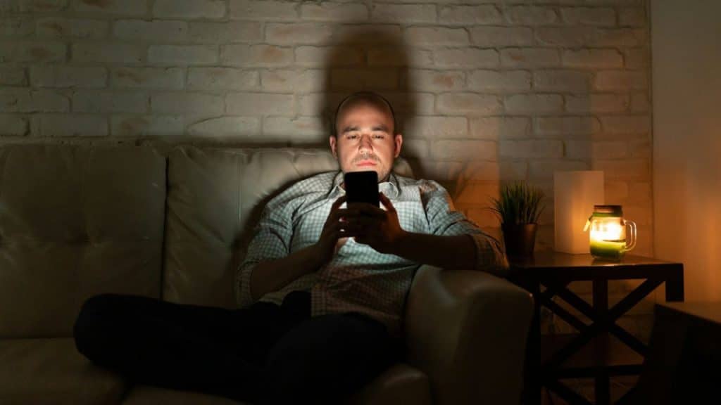 A man sits on a sofa in a dark room looking at a glowing smartphone.