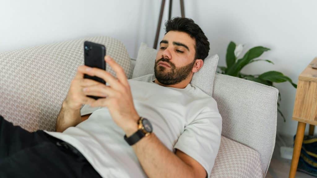 A bearded man lies on a couch while looking at a smartphone held in both hands.