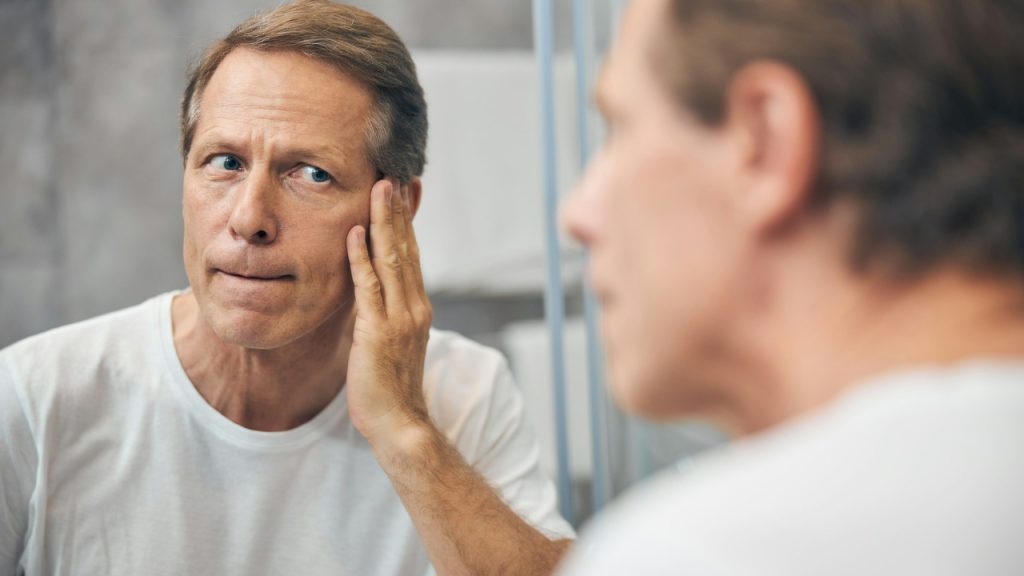 A middle-aged man touches his face while looking at his reflection in a mirror.