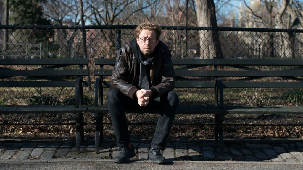A man in a leather jacket sits on a park bench with hands clasped.