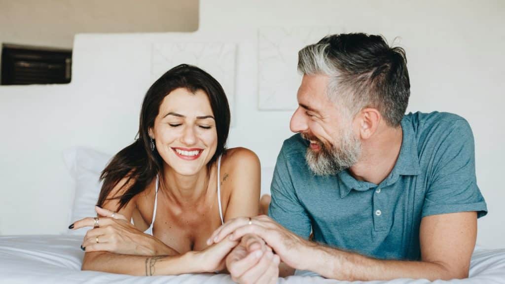 A man and woman lie on a bed smiling while holding hands and talking.