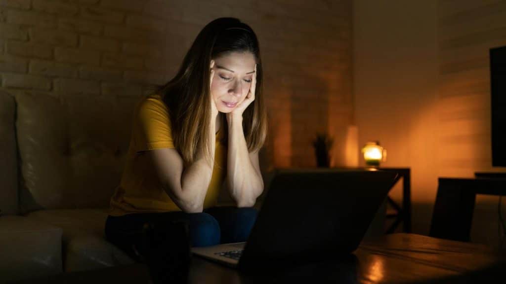 A woman in a yellow shirt looks stressed while sitting at a laptop in a dimly lit room.