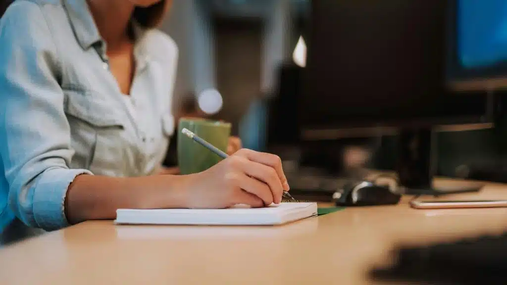 A woman writing in a notebook while holding a mug at her desk.