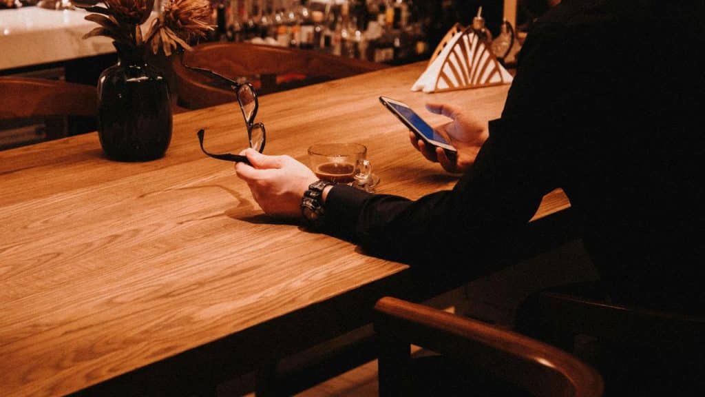 A person holding eyeglasses in one hand and a smartphone in the other at a wooden bar table