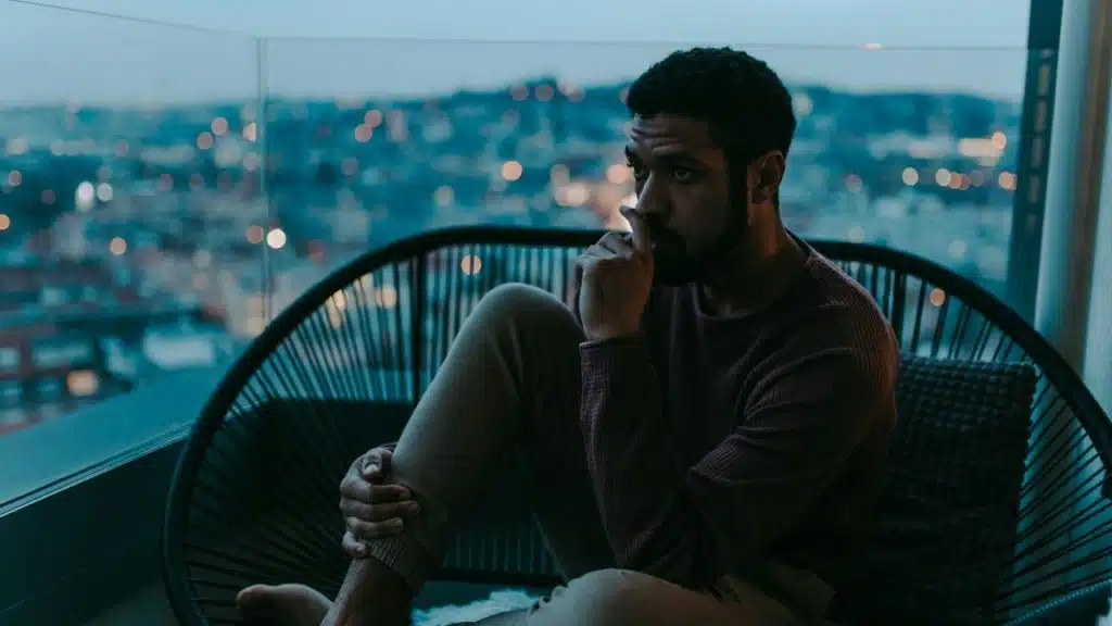 A man sits in a black chair on a balcony overlooking a city at twilight.
