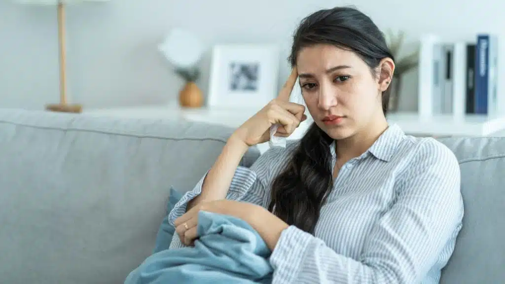 A young woman sits on a couch, holding her head, with a pensive expression.
