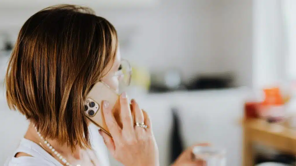 A woman wearing glasses talks on a smartphone while holding a cup indoors.