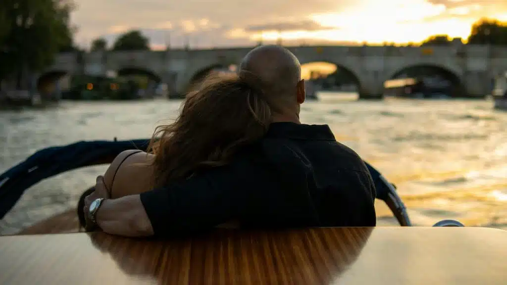A couple sitting close together on a boat, watching a river and a bridge at sunset.