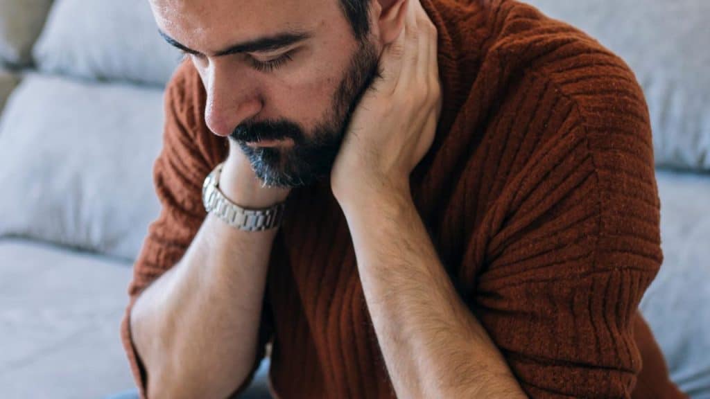 A bearded person sitting on a couch, holding their neck and looking down.