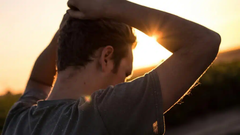 A man stands outdoors at sunset with his arms raised behind his head.