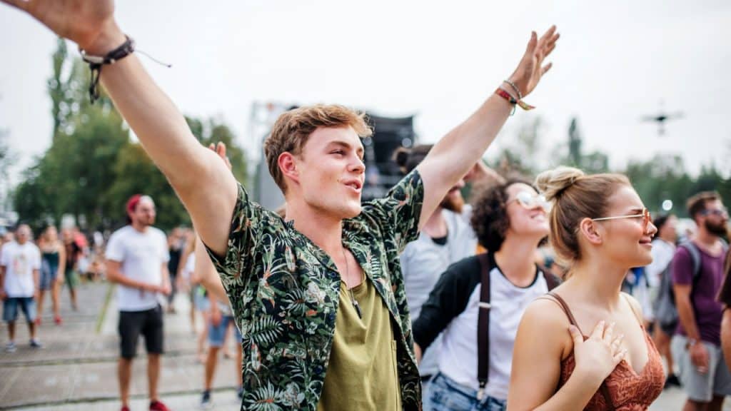 A young man with raised arms cheers alongside a crowd at an outdoor music festival.
