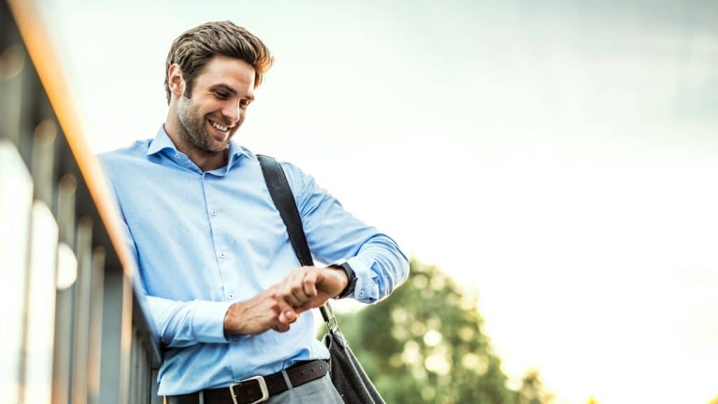 A man in a light blue shirt smiles while looking down at his wristwatch outdoors.