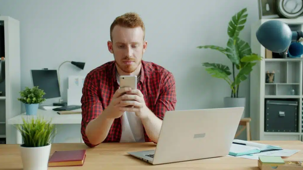 A man with red hair using a smartphone at a desk with a laptop.
