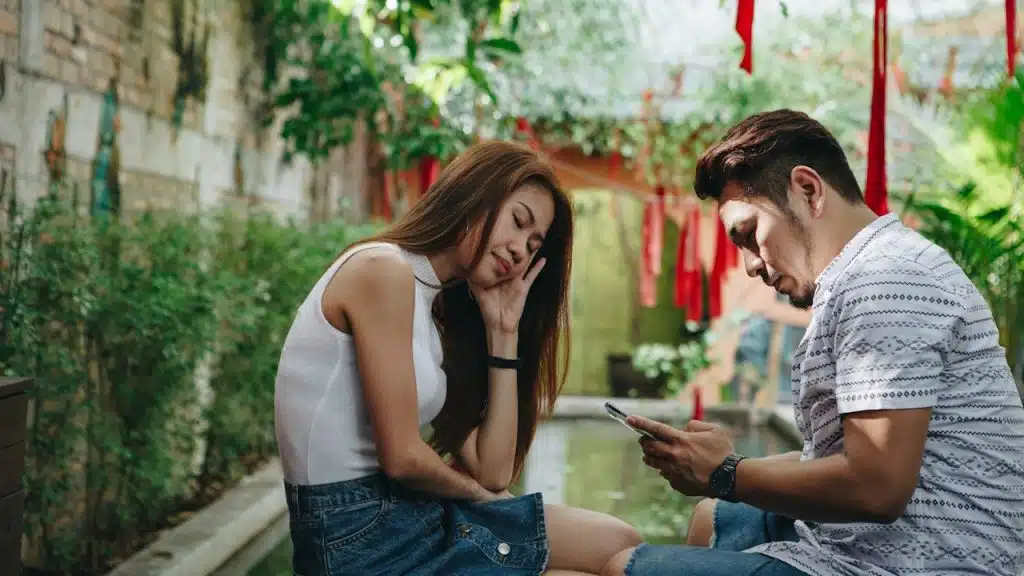 A woman looking exasperated and holding her cheek with her hand as a man texts while sitting in front of her.