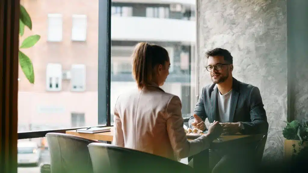 A man and a woman talking across a small table in a café by a window.