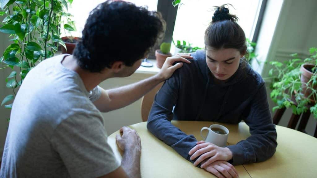 A man with a grey shirt comforts a woman in a black hoodie at a table.