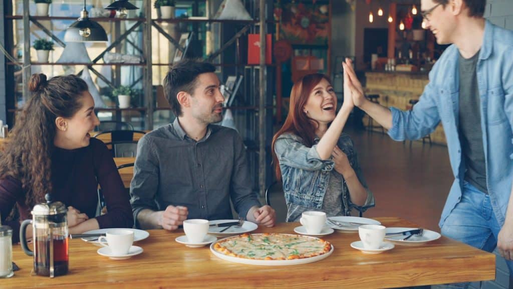 A group of friends having pizza at a cafe, where two people high-five each other.