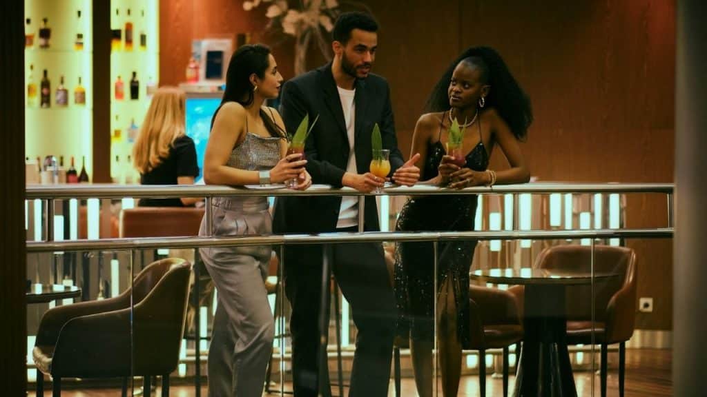 Three people stand at a glass railing in a dimly lit lounge holding colorful drinks.