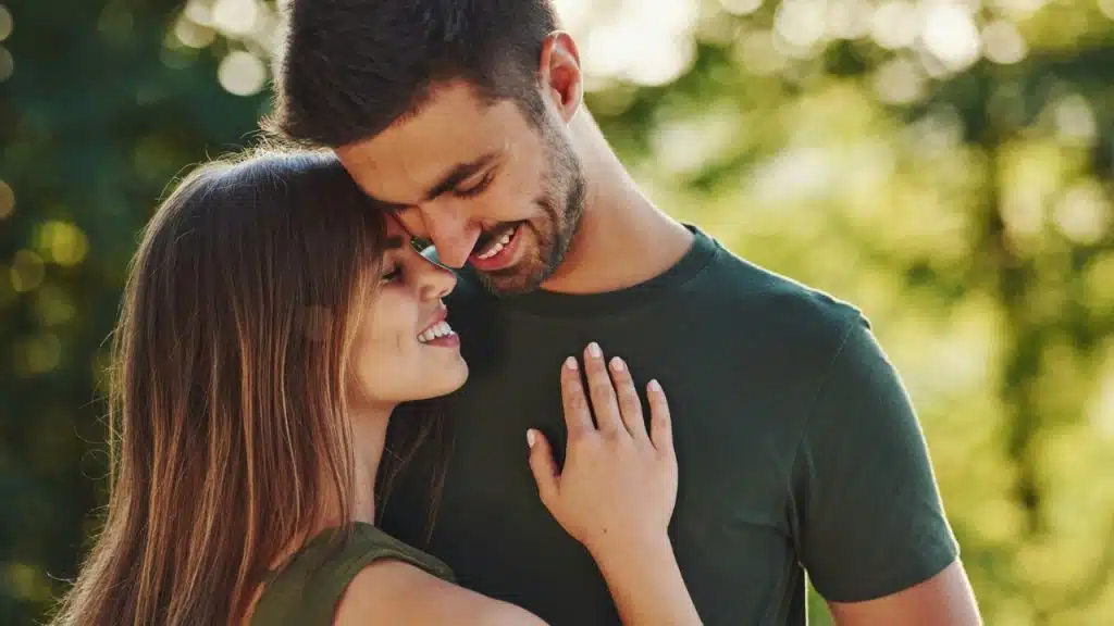 A smiling woman rests her hand on a man's chest while their heads touch outdoors.