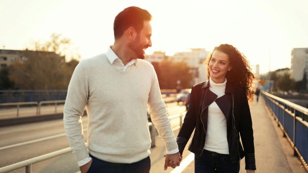 A man and woman hold hands and smile while walking outdoors at sunset.