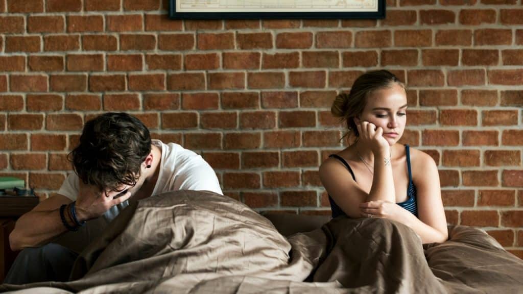 A man and woman sit back-to-back in bed with their heads down against a brick wall.