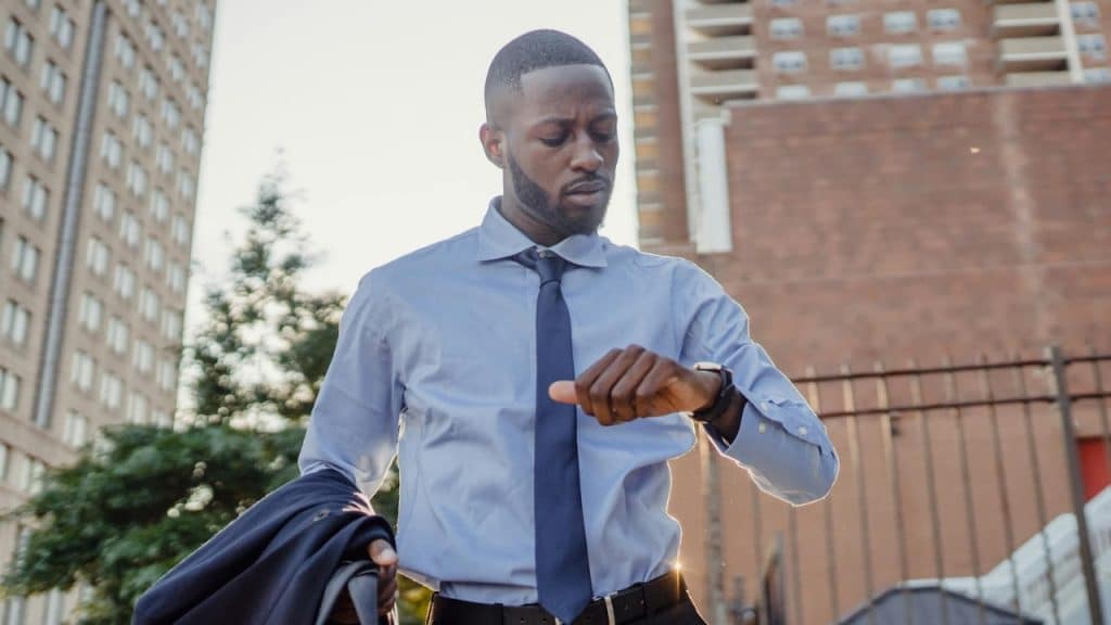 A man in a blue shirt and tie looks at his watch while holding a jacket.