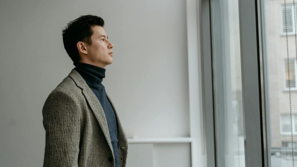 A man wearing a blazer and turtleneck looks out a large window in a room.