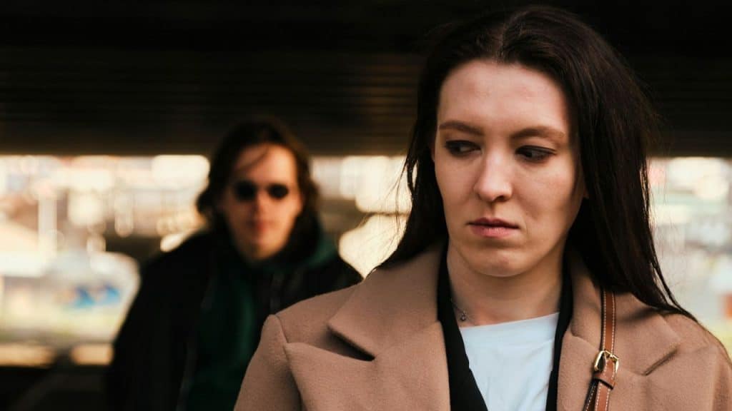 A woman in a tan coat looks downward while a blurred person stands behind her.