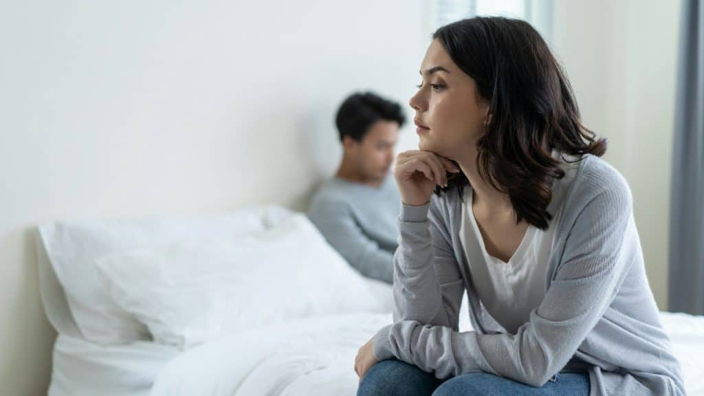A woman sits on the edge of a bed with a hand on her chin.